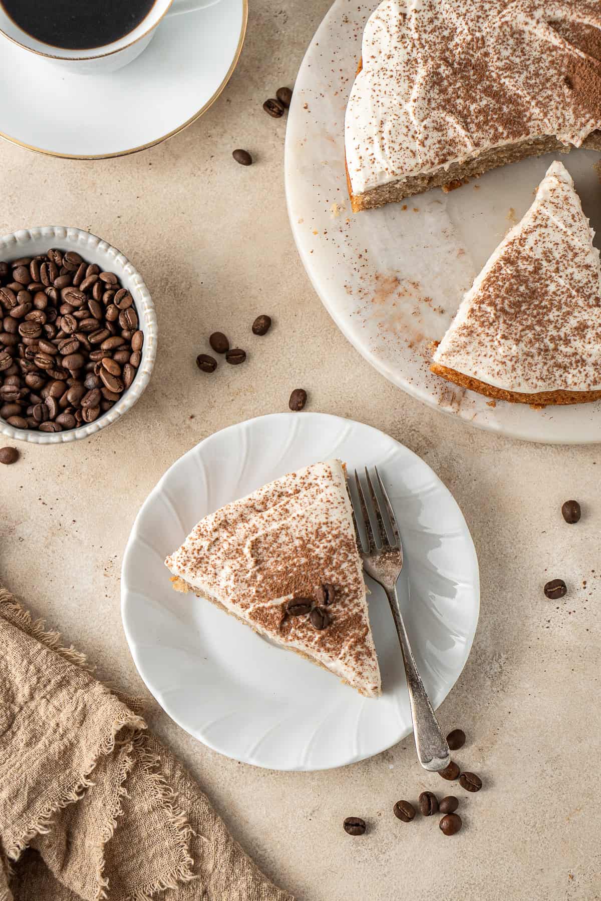 A slice of cappuccino cake served on a plate with a fork with coffee beans around it.