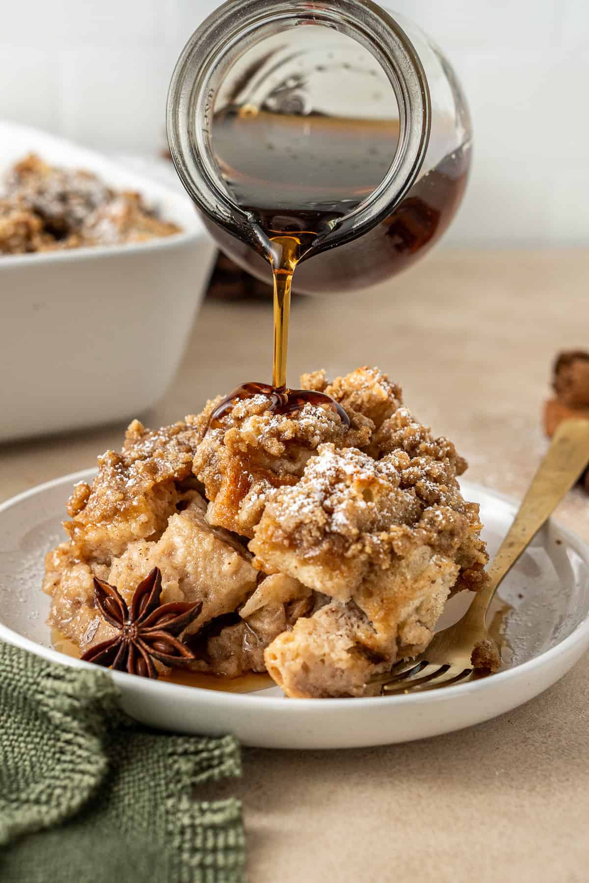 A jug of maple syrup being poured over a slice of French toast casserole.