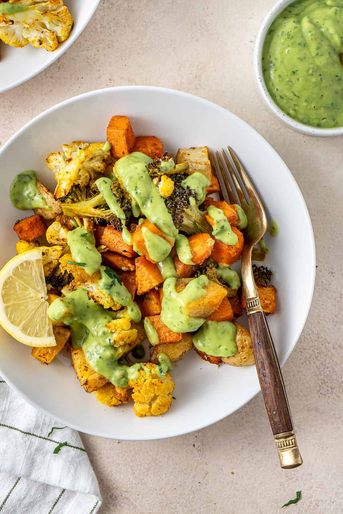 Close up of a roasted vegetable bowl with green goddess sauce, lemon and a fork.
