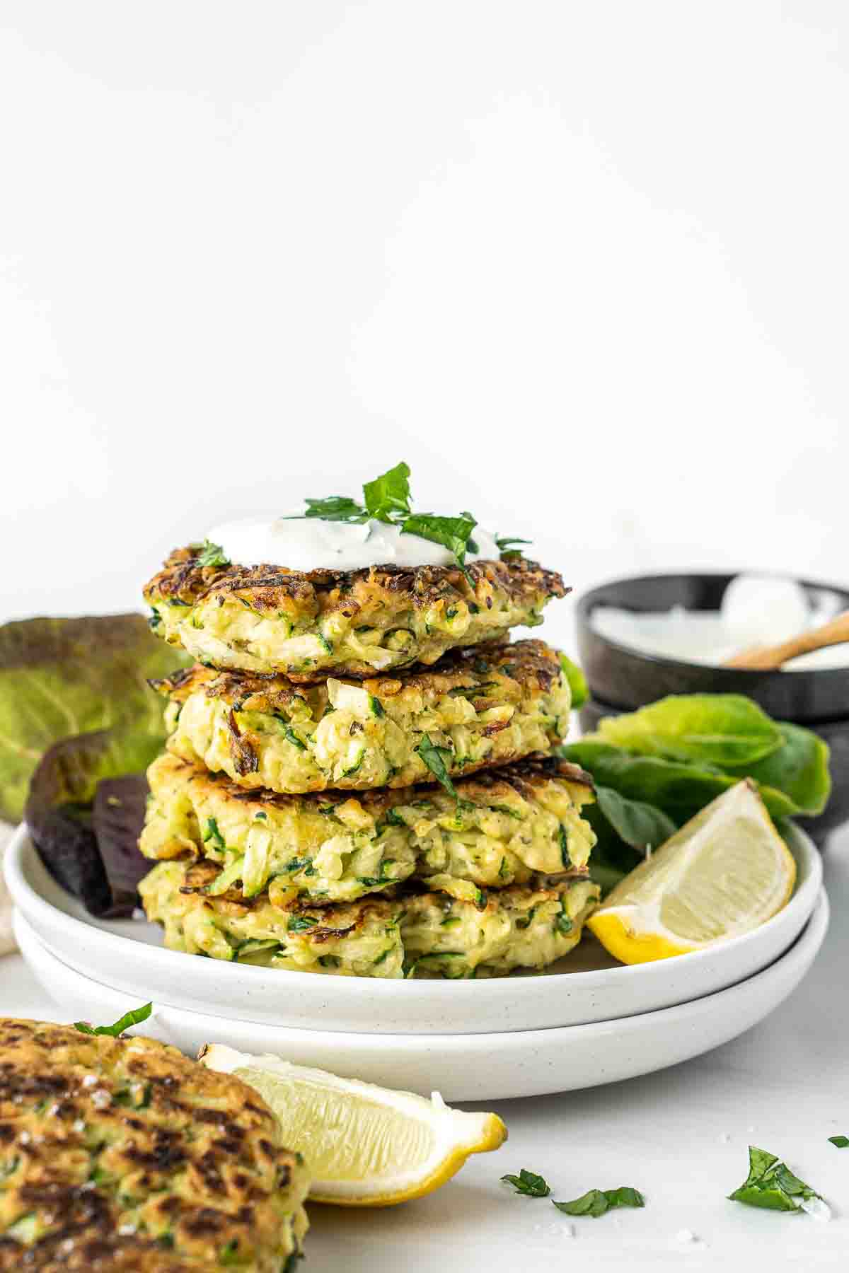 Stack of 4 zucchini fritters with sour cream on a plate with lemon.