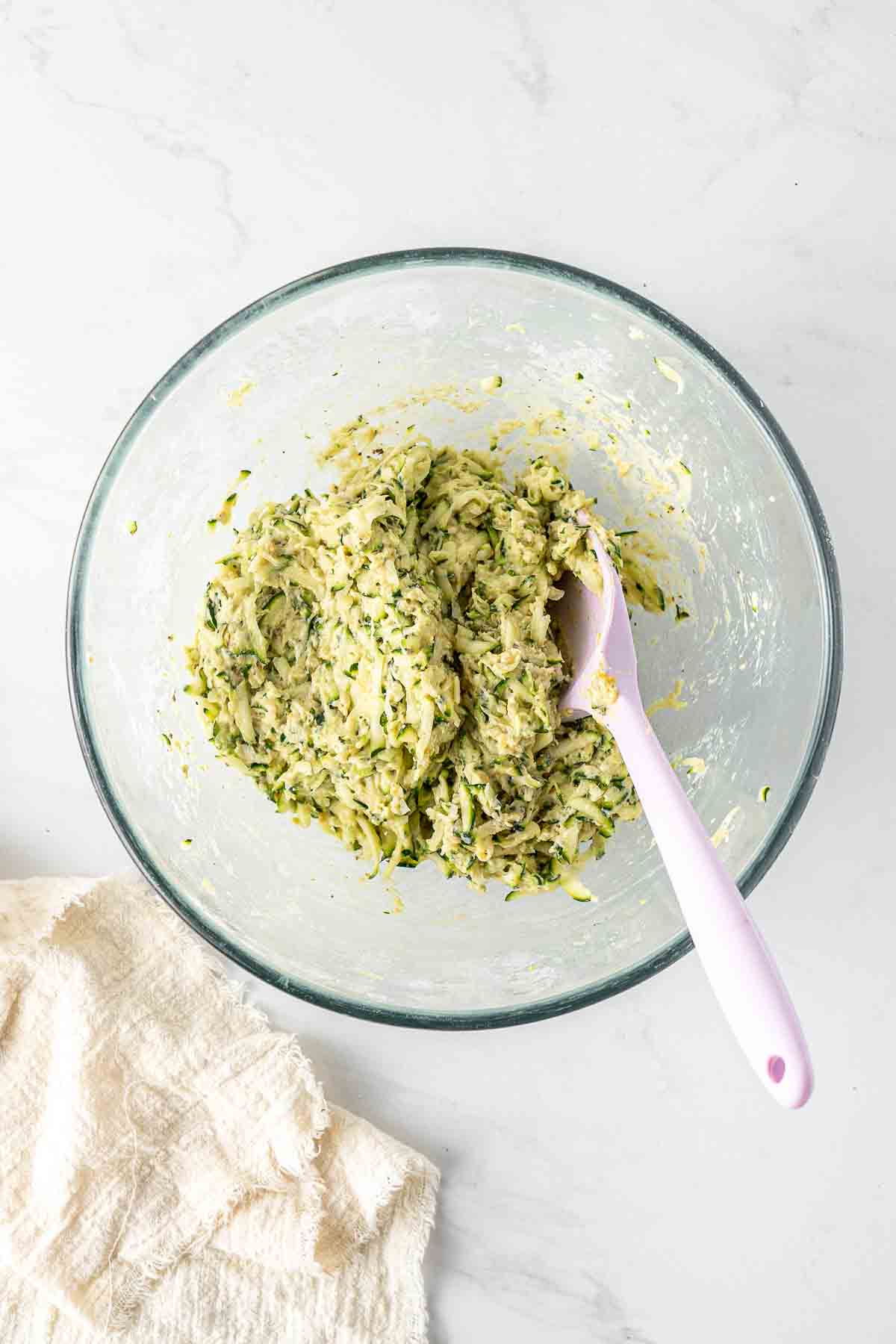 Zucchini fritter batter in a large glass bowl with a spatula. 