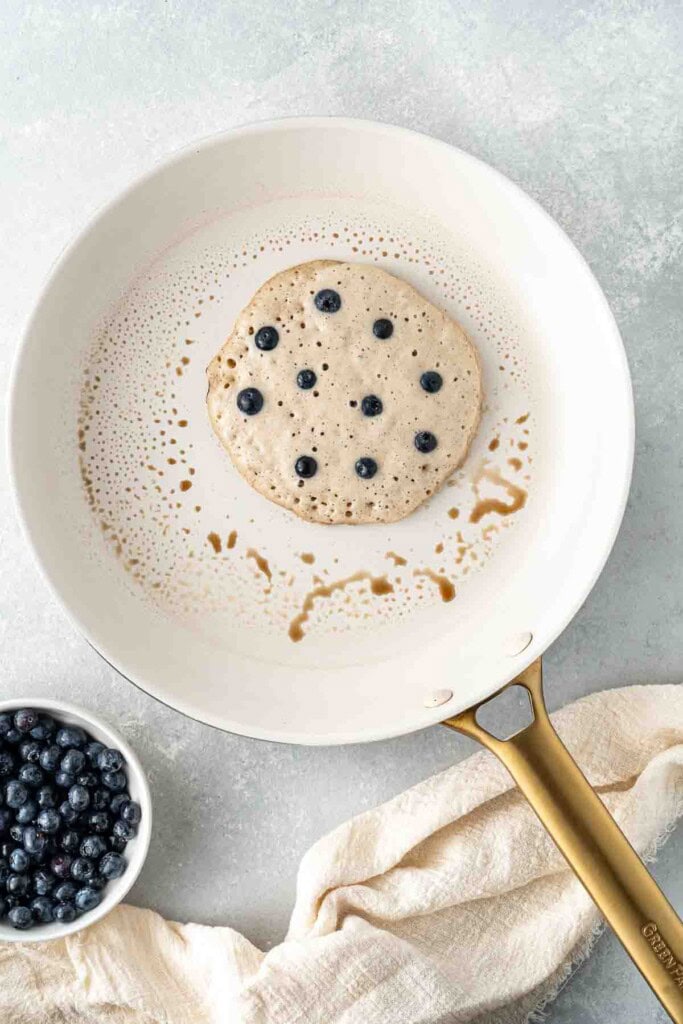 Adding blueberries to the pancake in the frying pan.
