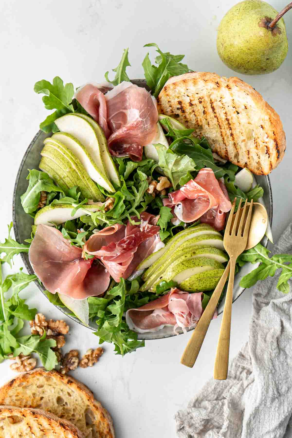 Pear and prosciutto salad in a bowl with toasted bread and gold cutlery. 