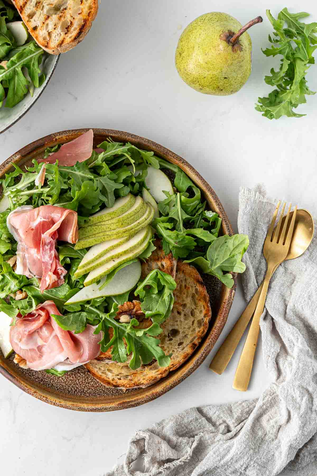 Pear and prosciutto salad on a plate with toasted bread and gold cutlery. 