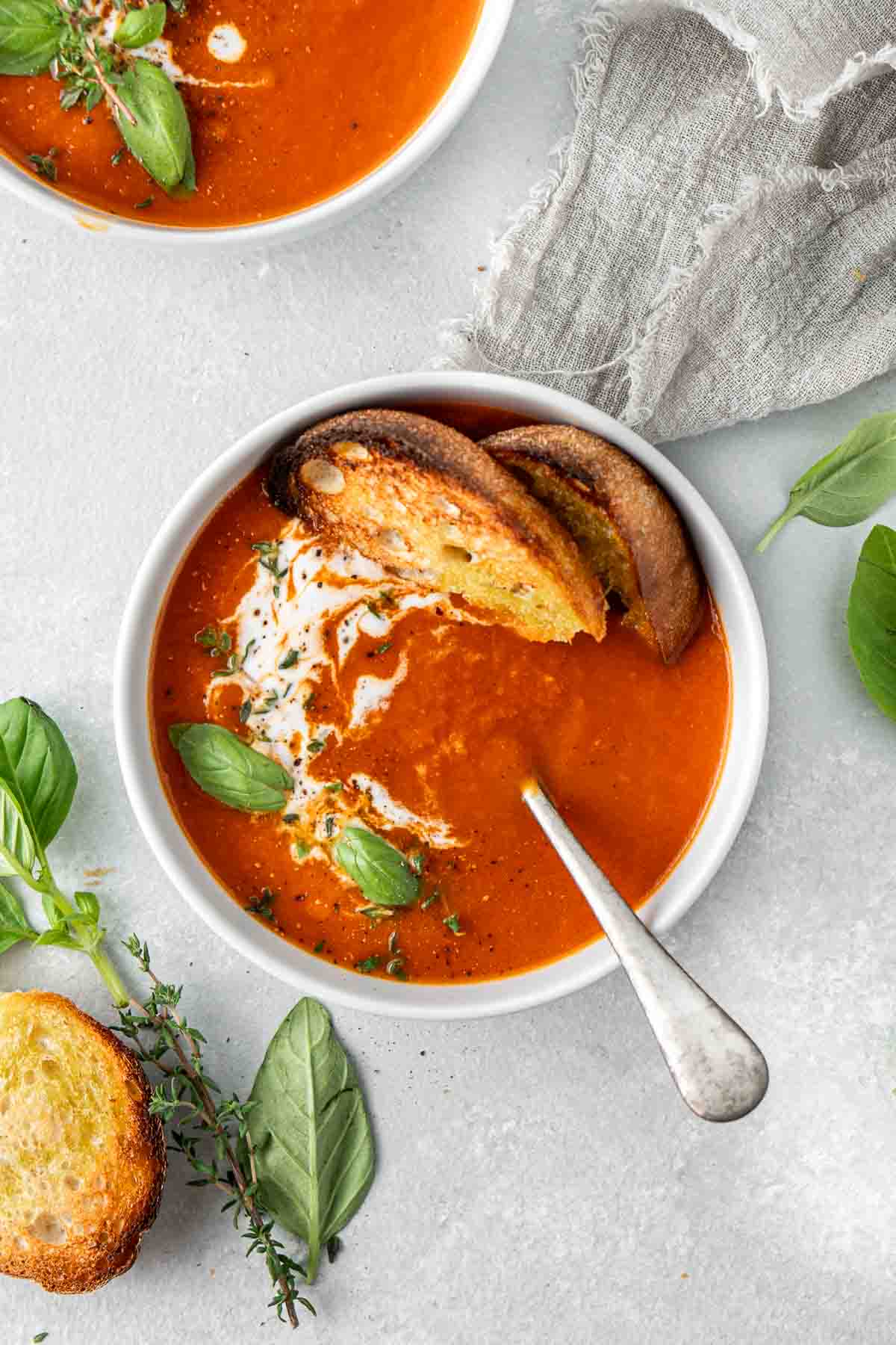 Overhead shot of a bowl of tomato soup with toasted bread and a spoon garnished with herbs.