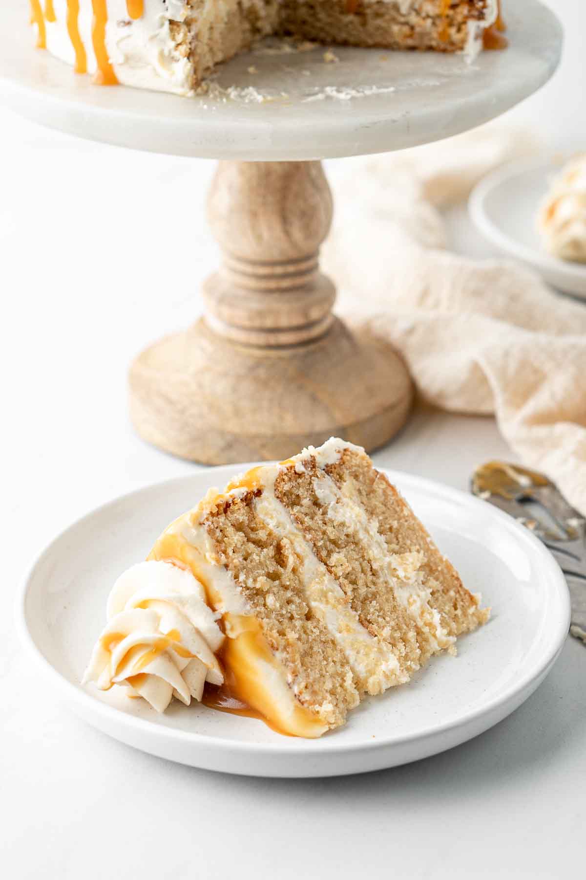 Slice of caramel cake on a white plate with the cake stand in the background.
