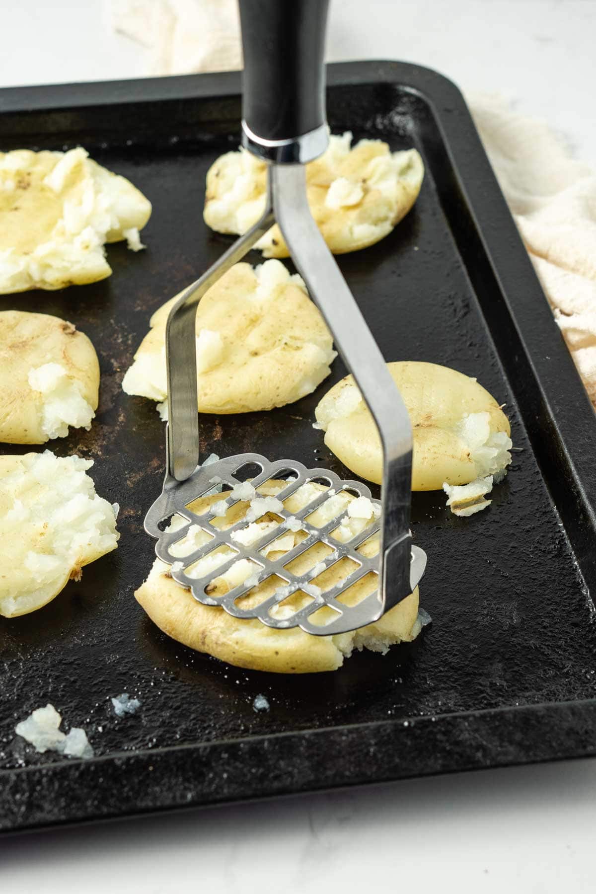 Potatoes on a baking tray being smashed with a potato masher.
