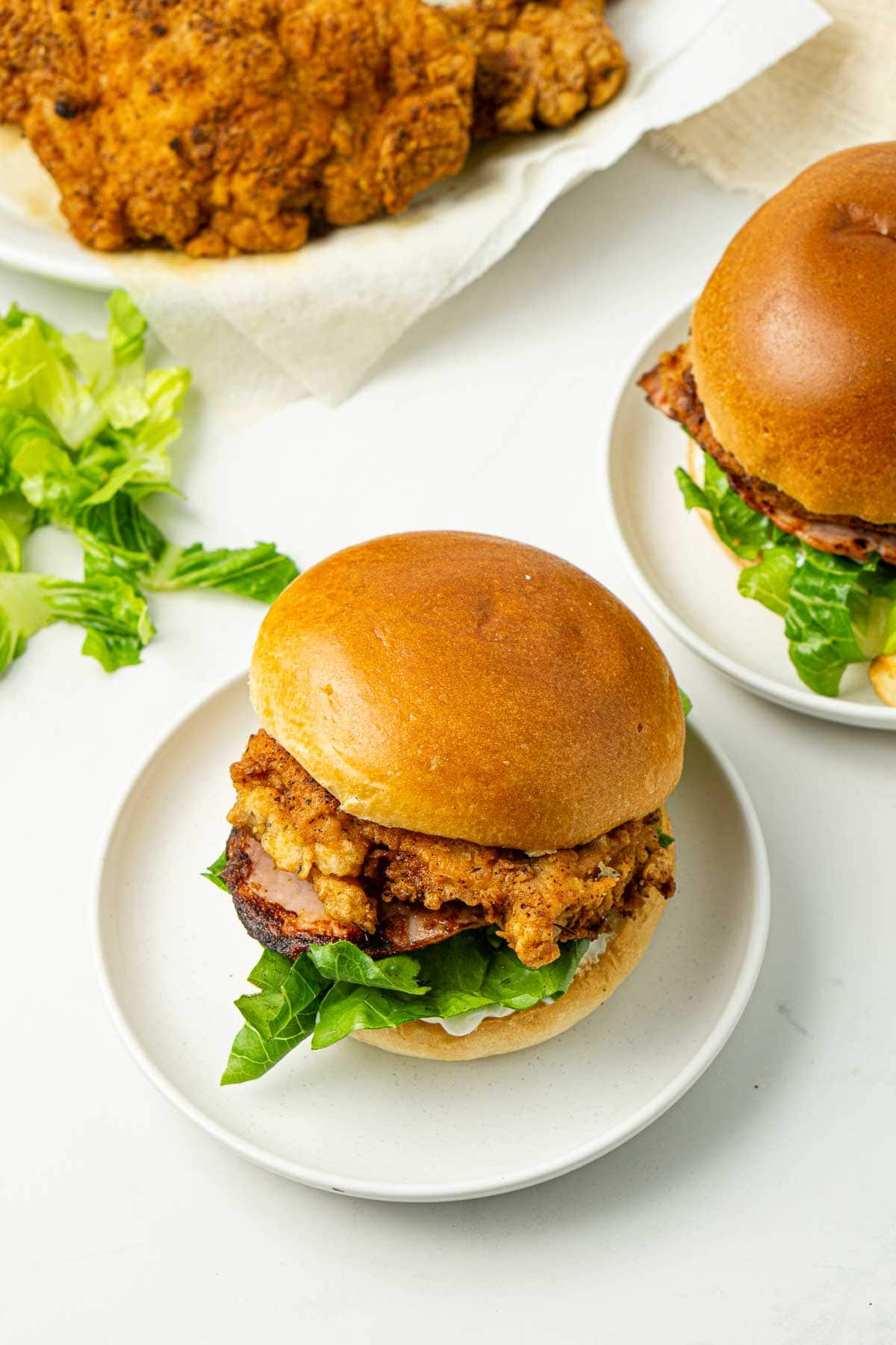 Two crispy chicken burgers on white plates with fried chicken in the background.