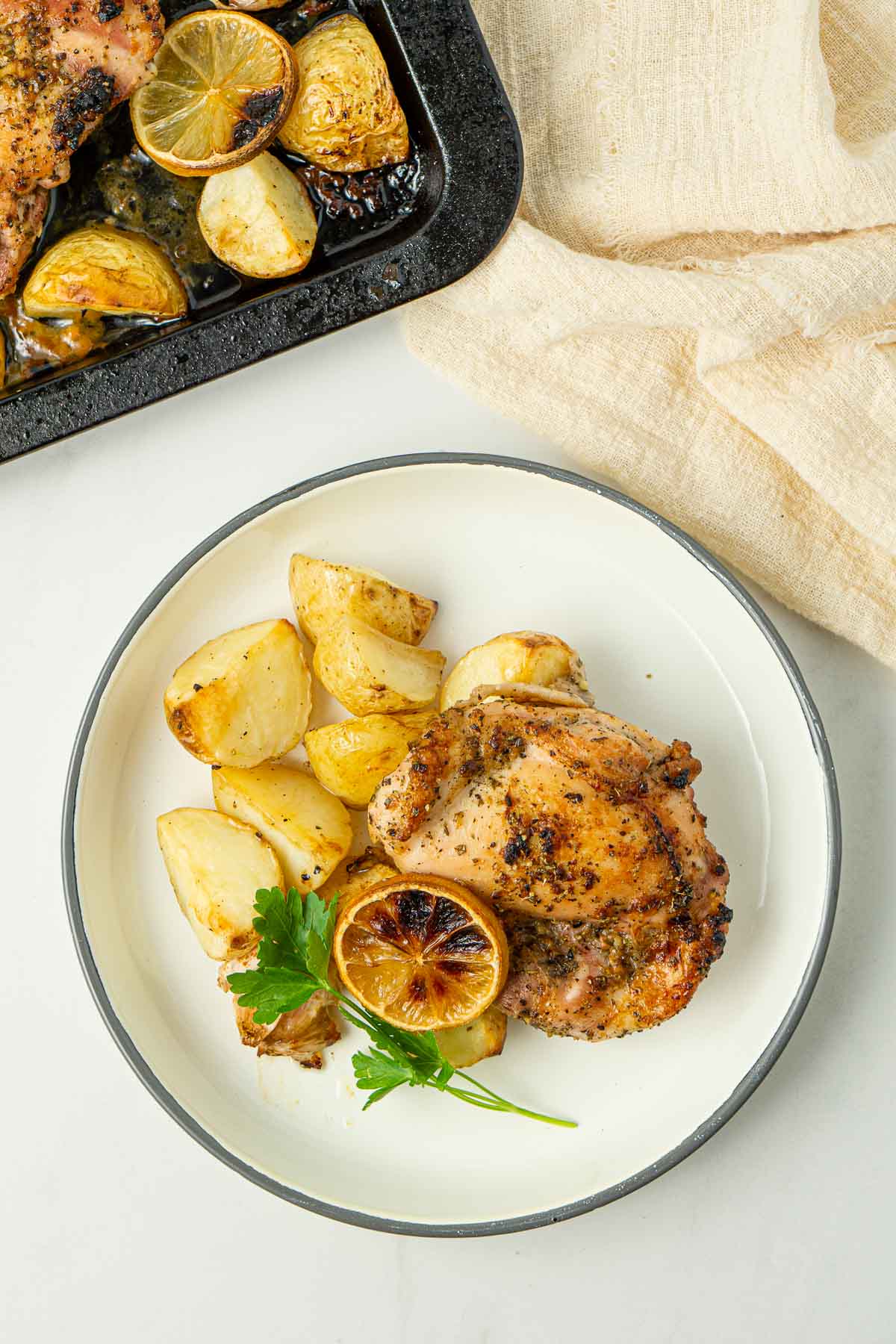 Chicken, potatoes, lemon and parsley on a white plate with a grey rim.