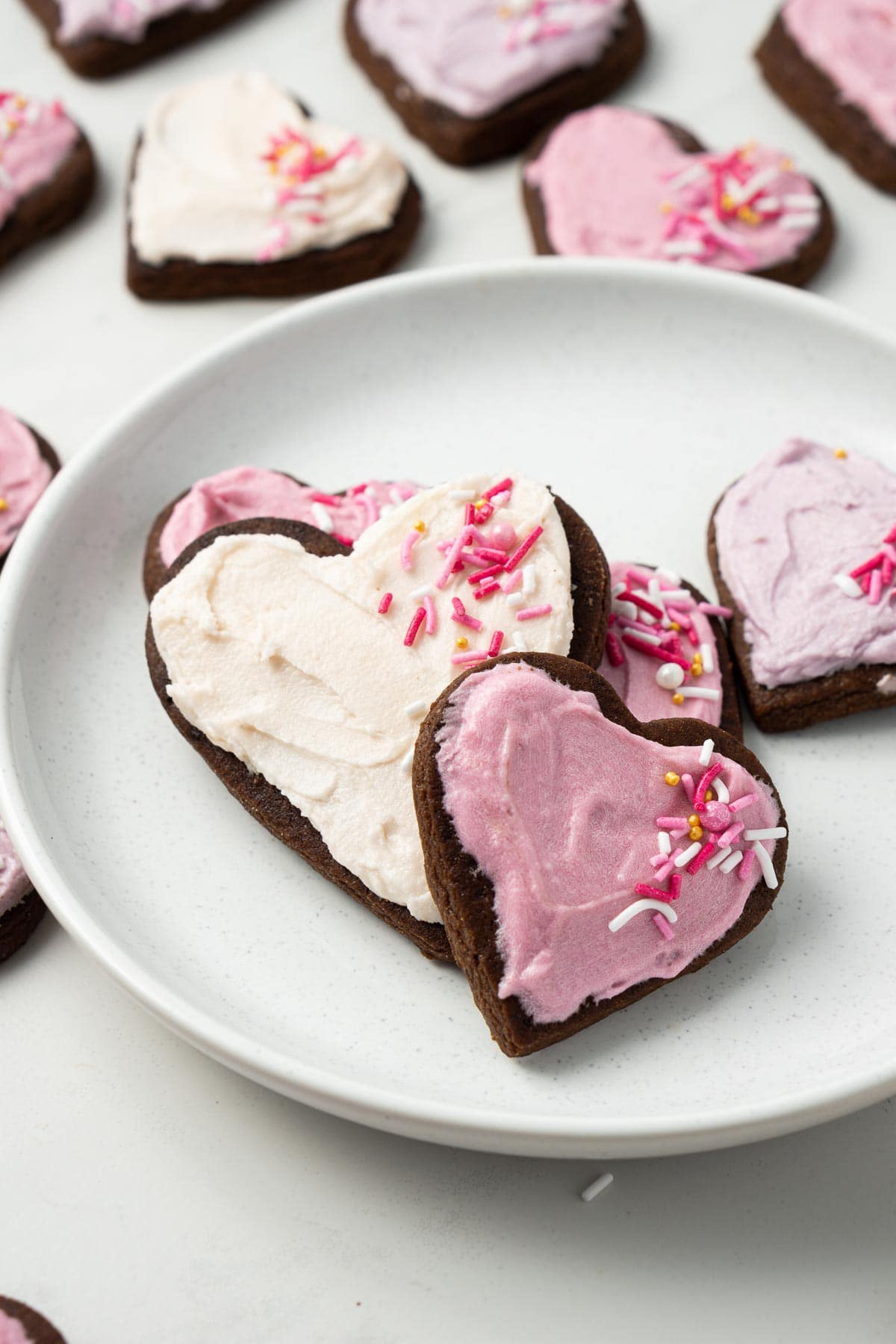 Close up of pink and purple iced chocolate sugar cookies laid out on a small white plate with more cookies around the outside.