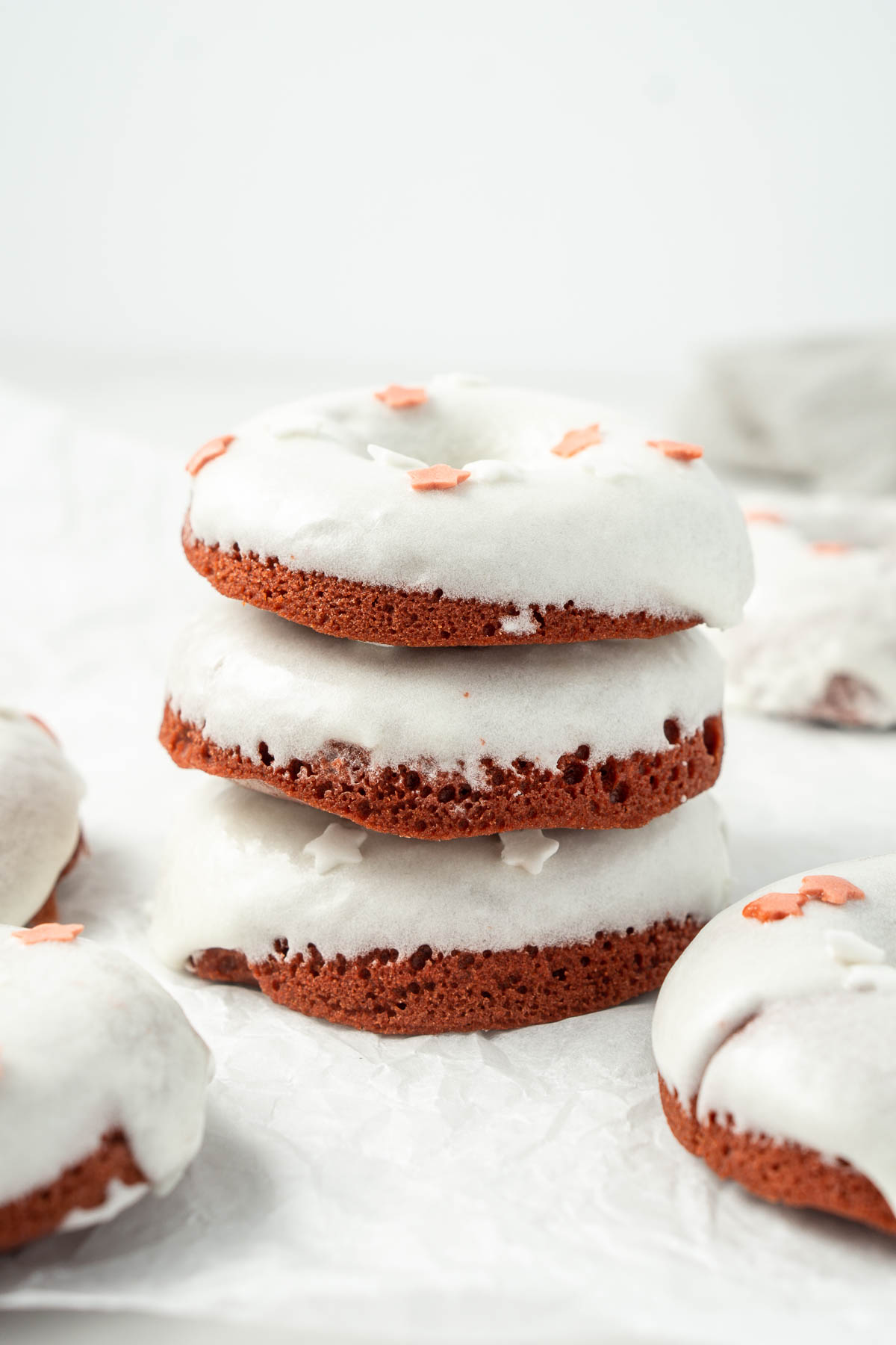 Three red velvet doughnuts staked on top of each other with white cream cheese glaze and red and white sprinkles.
