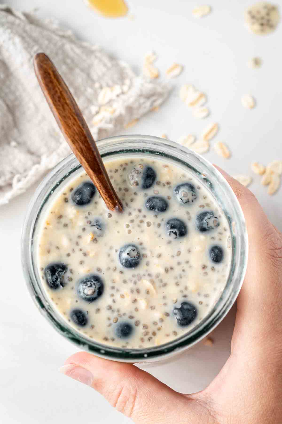 Hand holding vegan overnight oats with a spoon and blueberries.