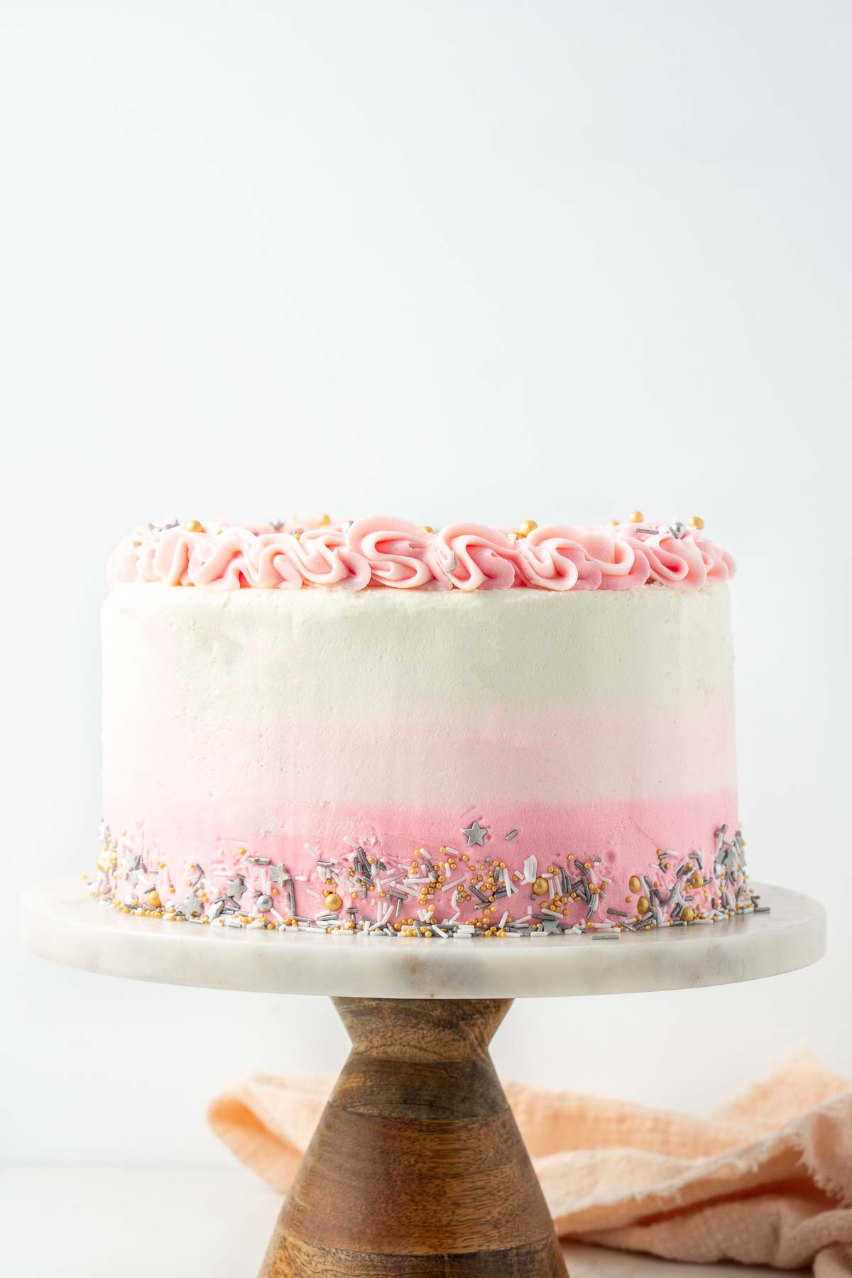 Vegan vanilla cake with pink ombre frosting on a marble and wood cake stand.