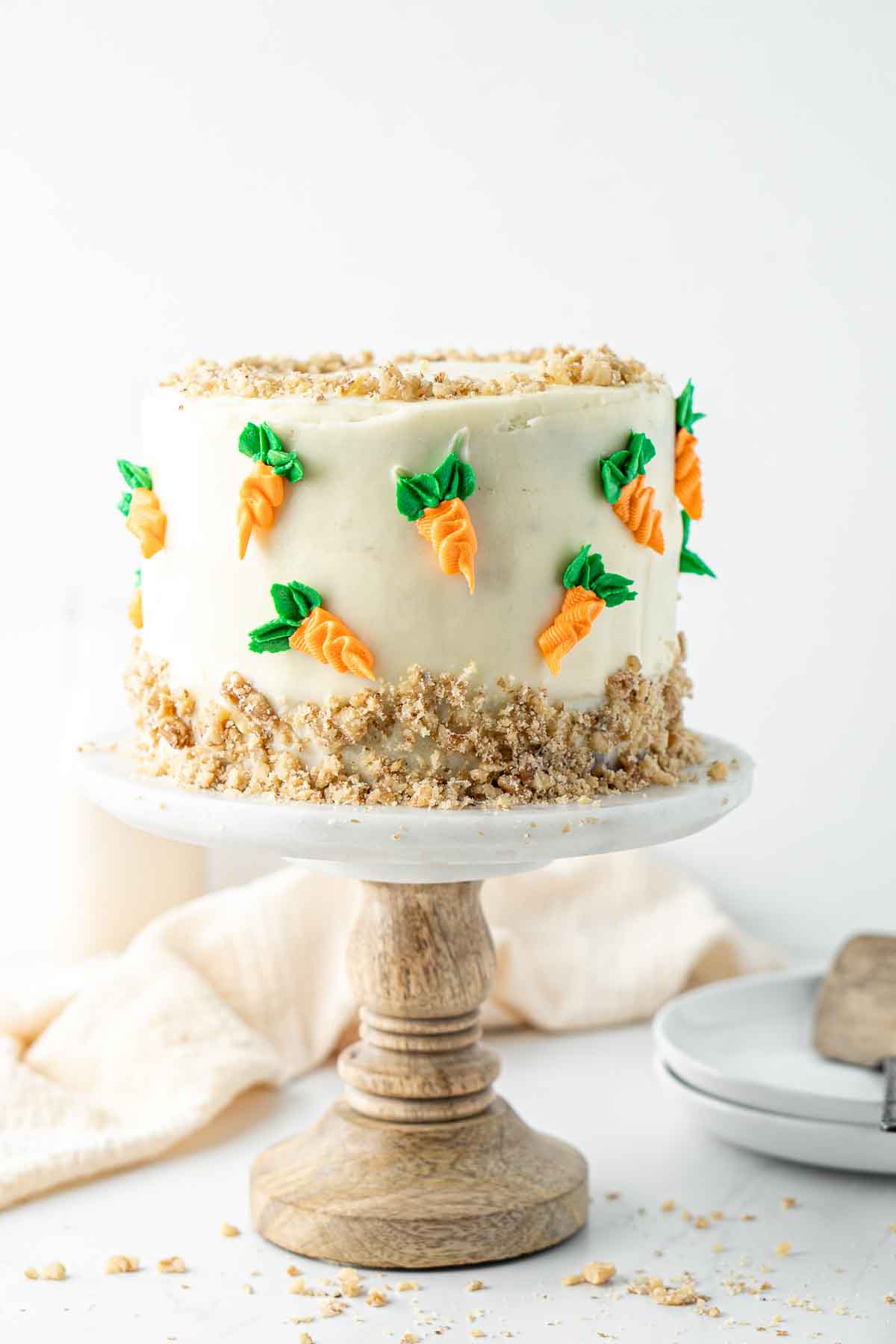 Vegan carrot cake on a cake stand decorated with cream cheese frosting, crushed walnuts and buttercream carrots.