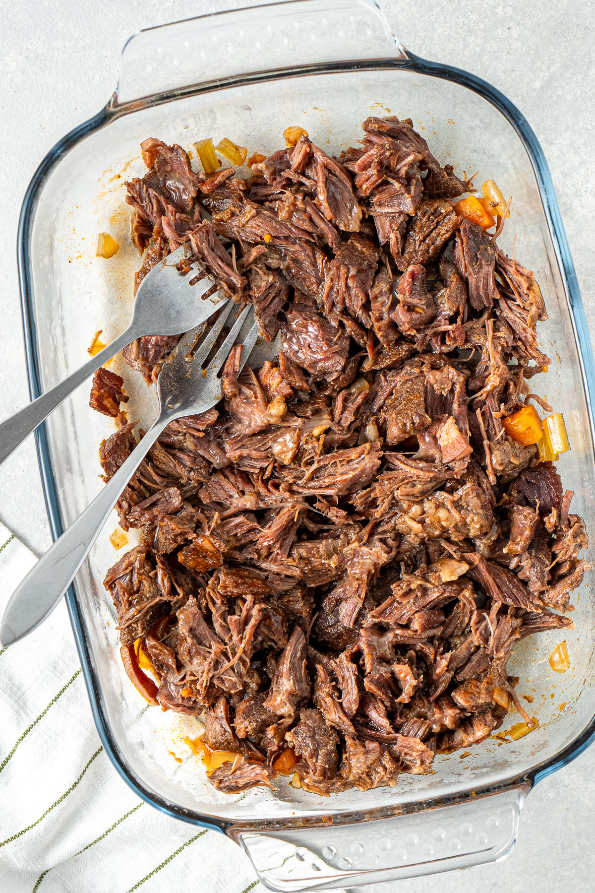 The beef cheeks shredded with two forks in a glass dish.