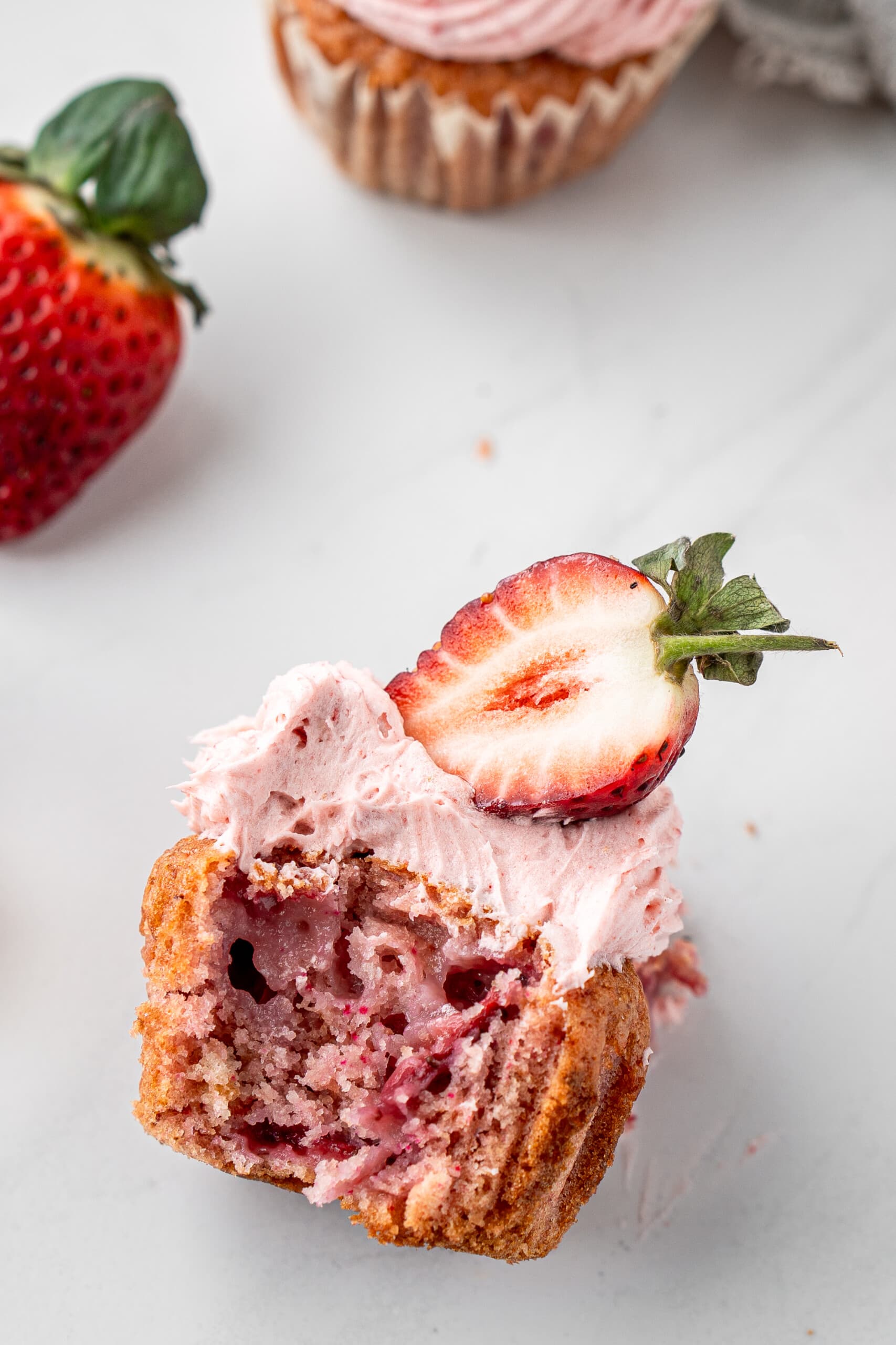 Close up of a strawberry cupcake with a bite taken and a fresh strawberry on top.