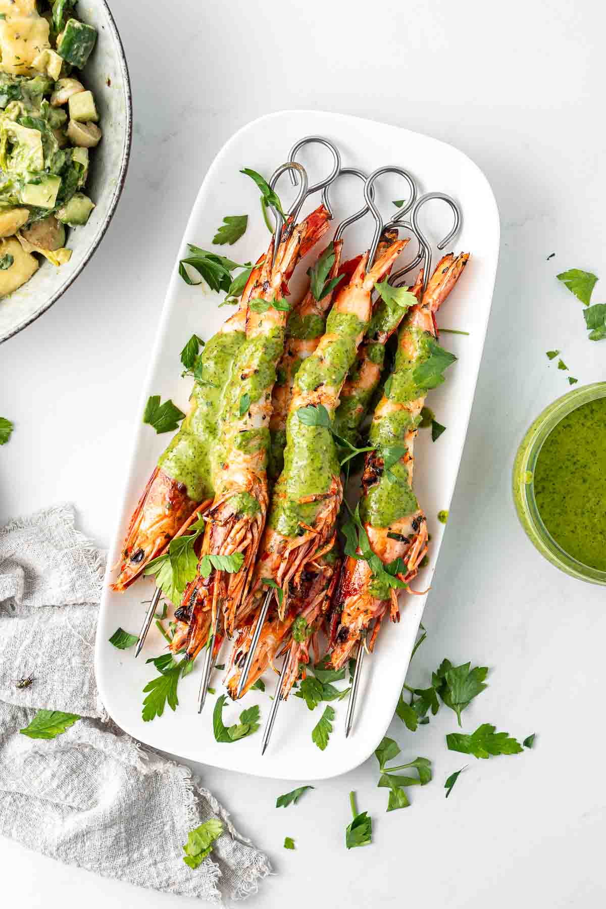 Grilled prawns on skewers on a white serving plate with green sauce and parsley.