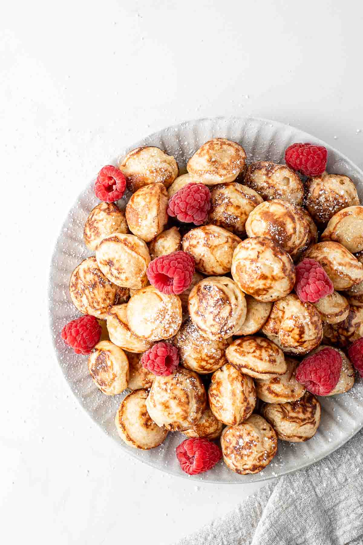 Pile of mini dutch pancakes with raspberries and icing sugar on a plate.