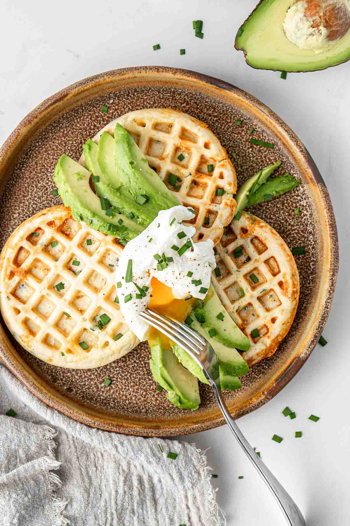 Overhead of poached egg and avocado on savoury waffles with fresh chives and a fork.