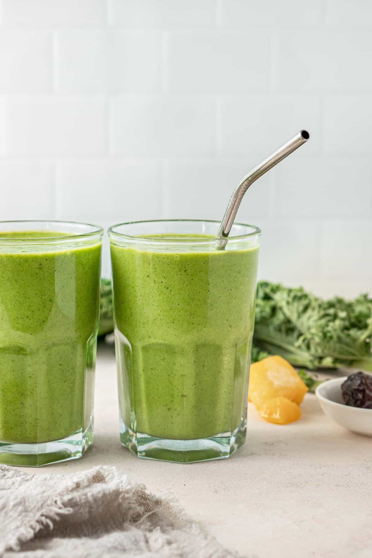 Two green smoothies in glasses, one with a metal straw.