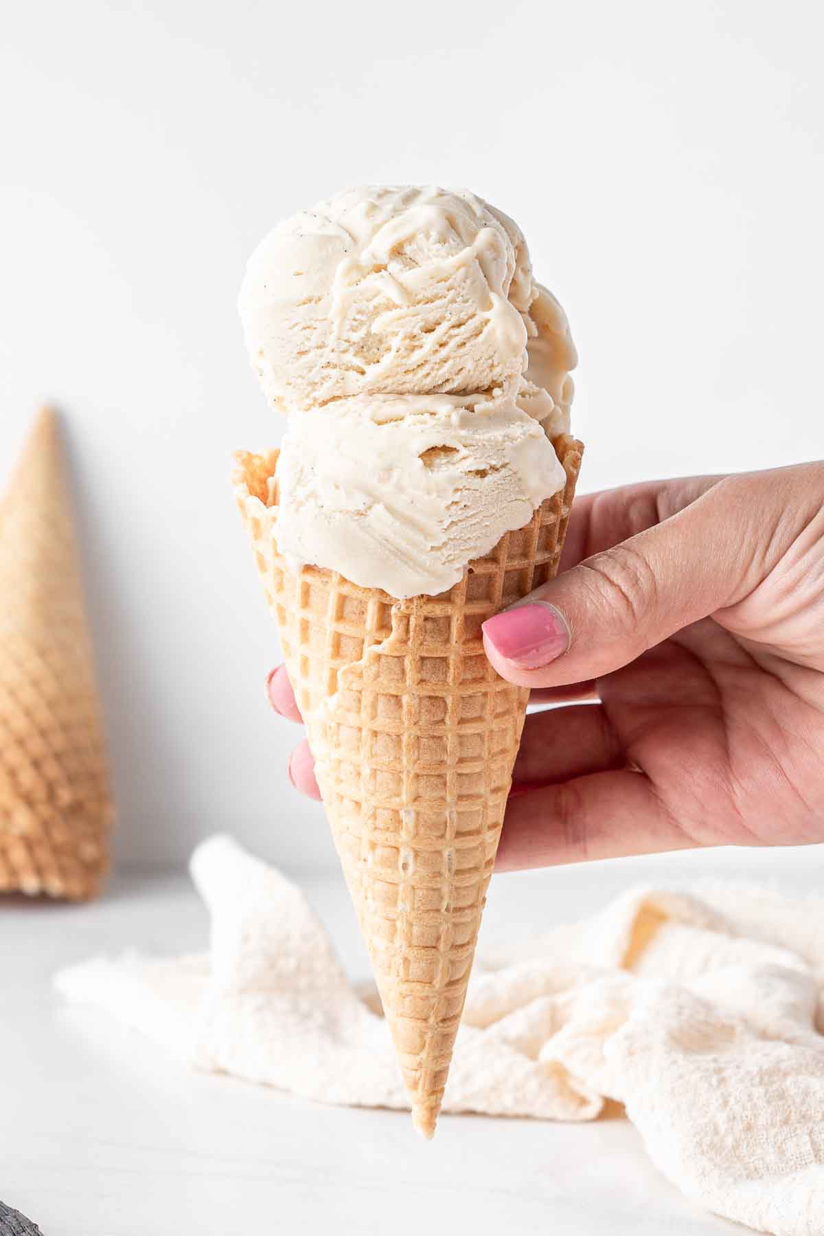 A hand holding an ice cream cone with two scoops of vegan vanilla ice cream.