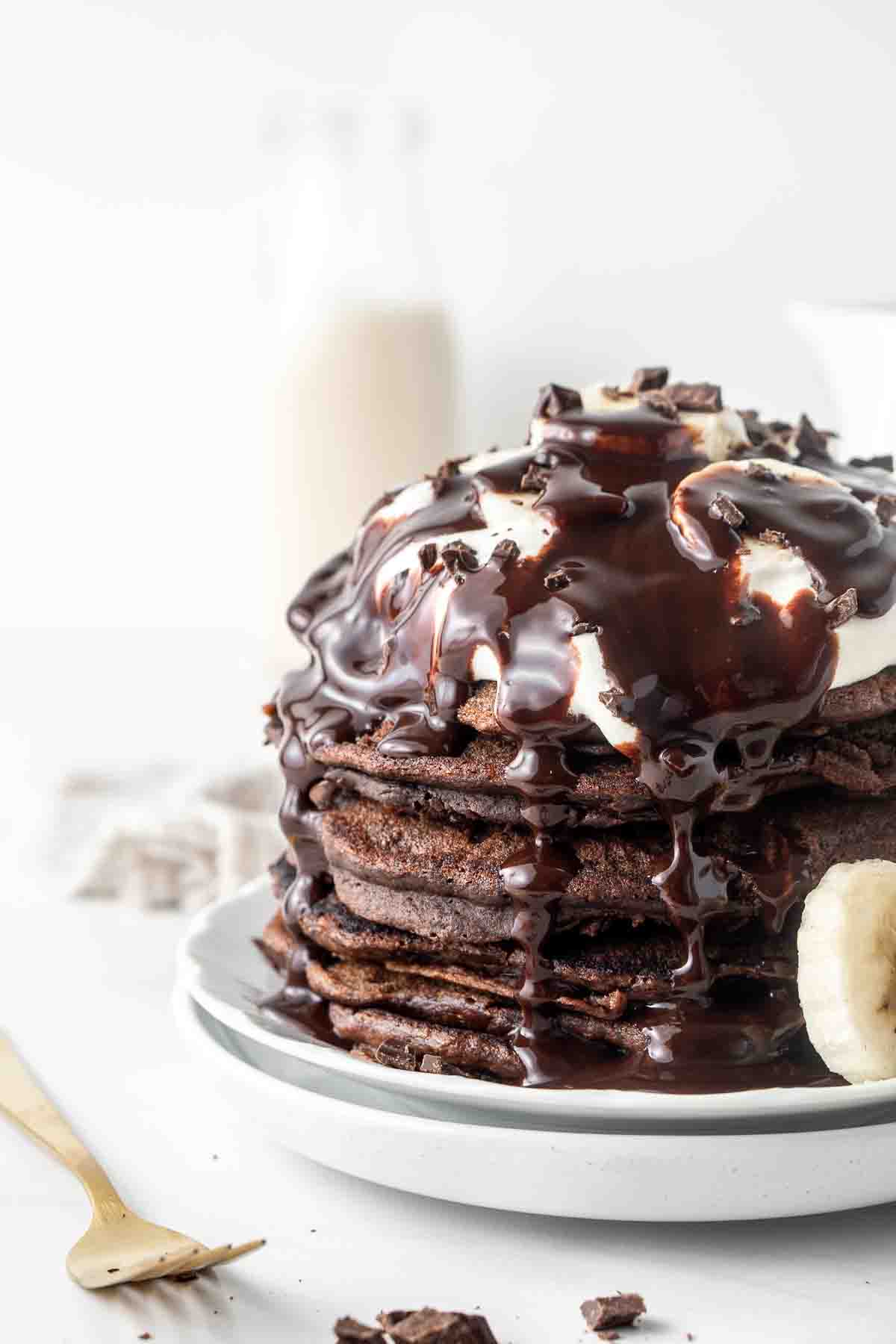 Close up of stack of chocolate pancakes on a white plate covered in chocolate syrup.