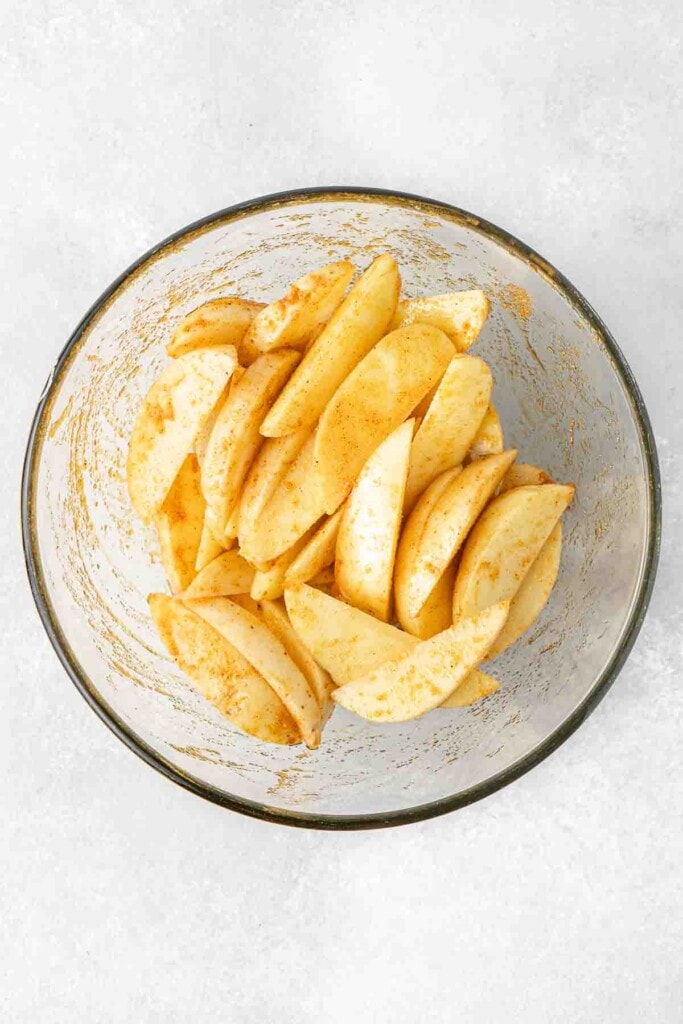 Potato wedges being tossed in oil and spiced in a glass bowl.