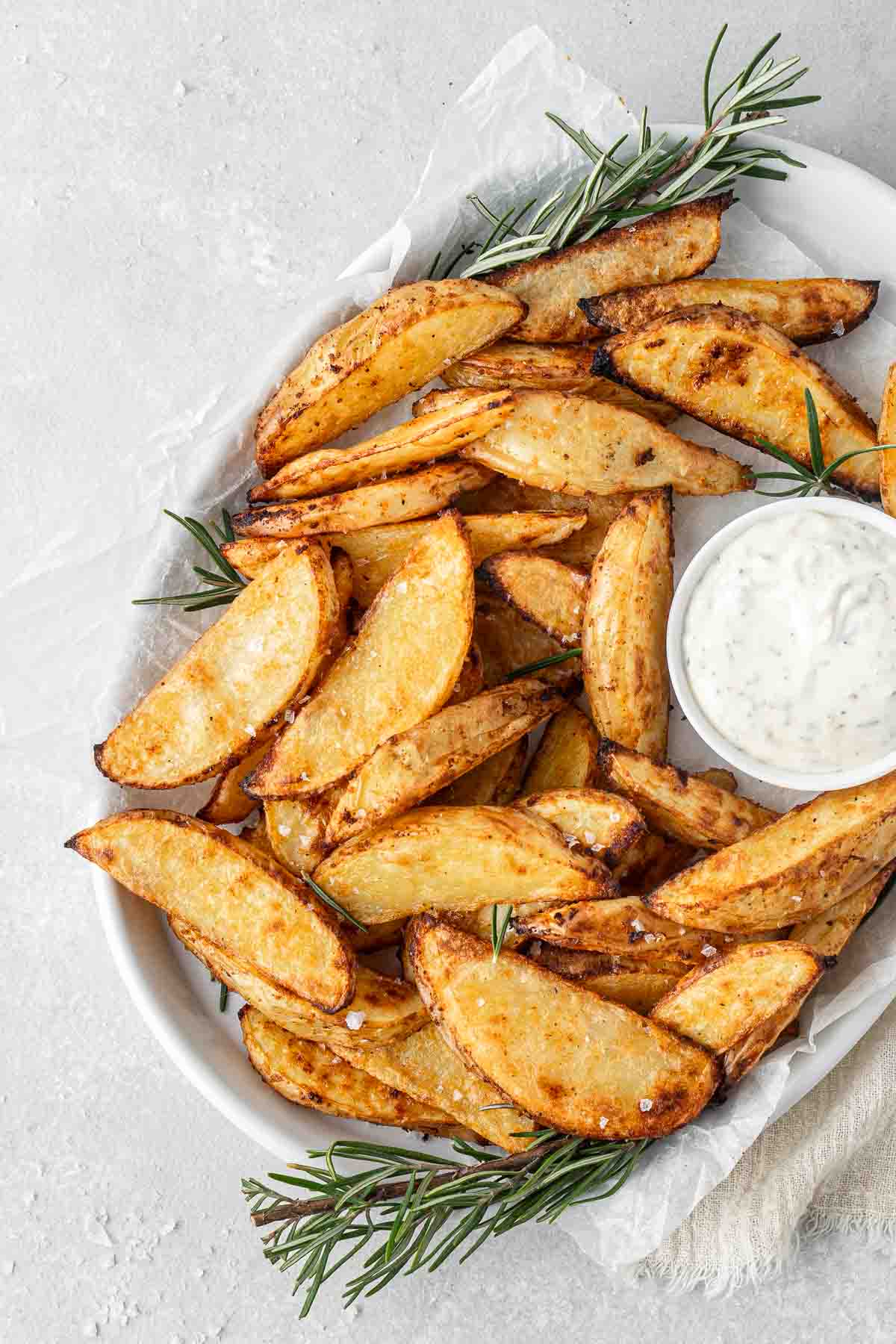 Close up of potato wedges on a serving platter with herb mayo and rosemary.