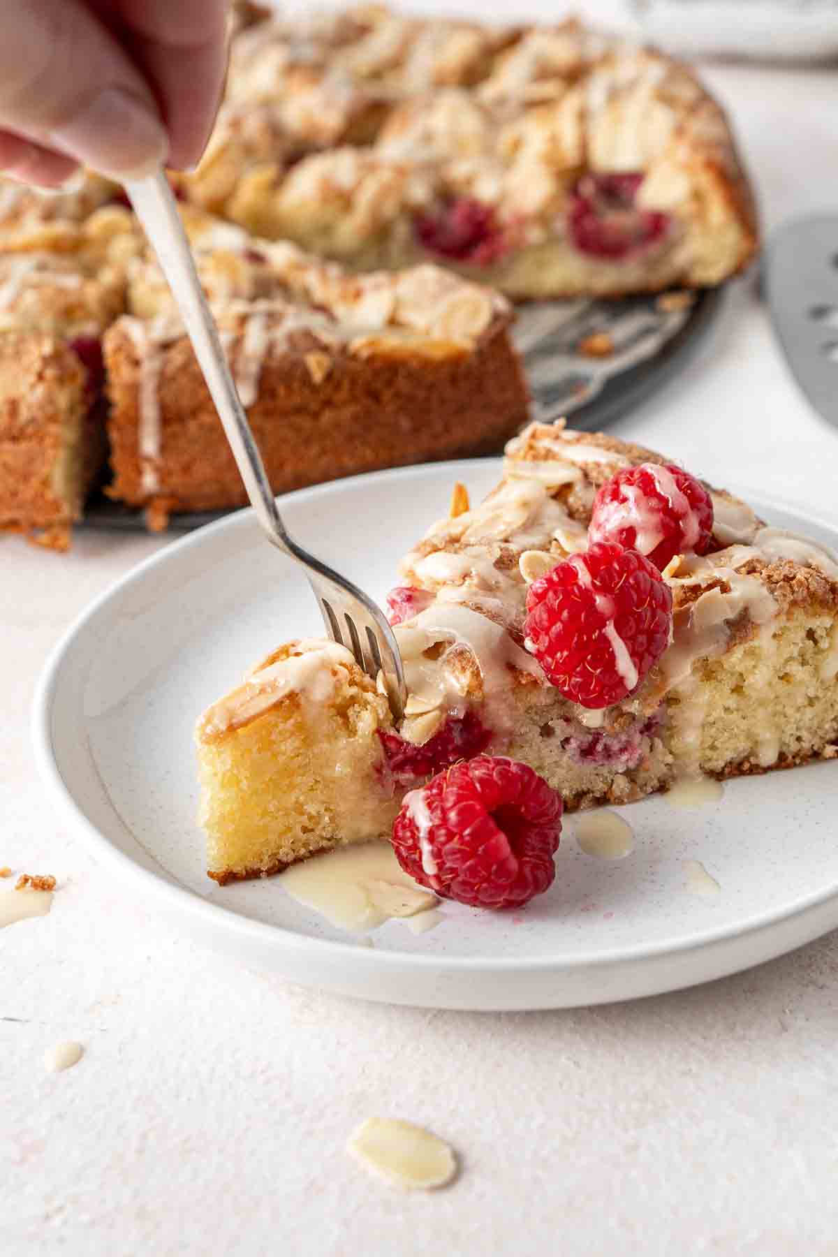 Close up of a fork taking a bite of a slice of almond and raspberry cake on a plate.
