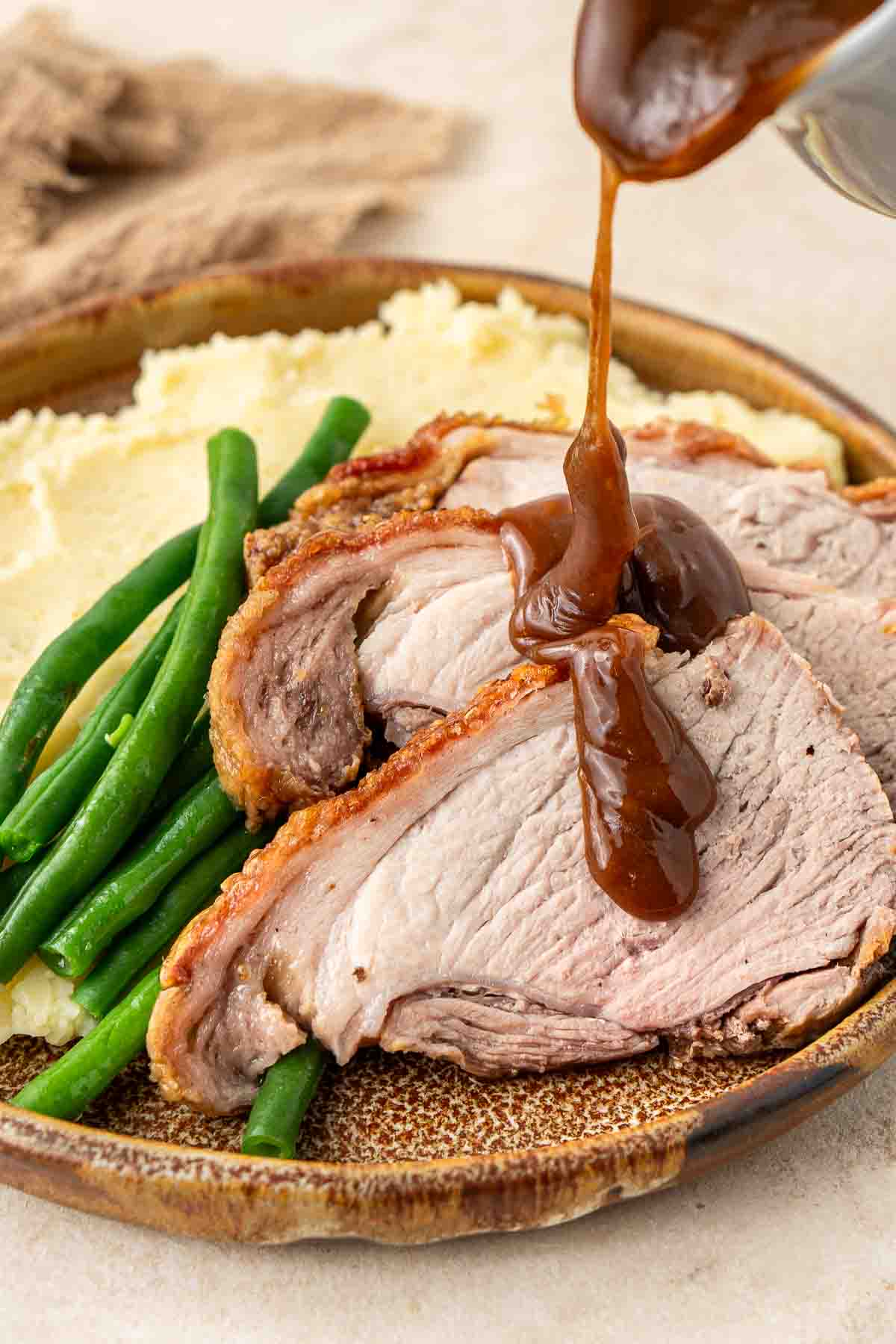 Gravy being drizzled over pork roast slices on a plate with green beans and mashed potato.
