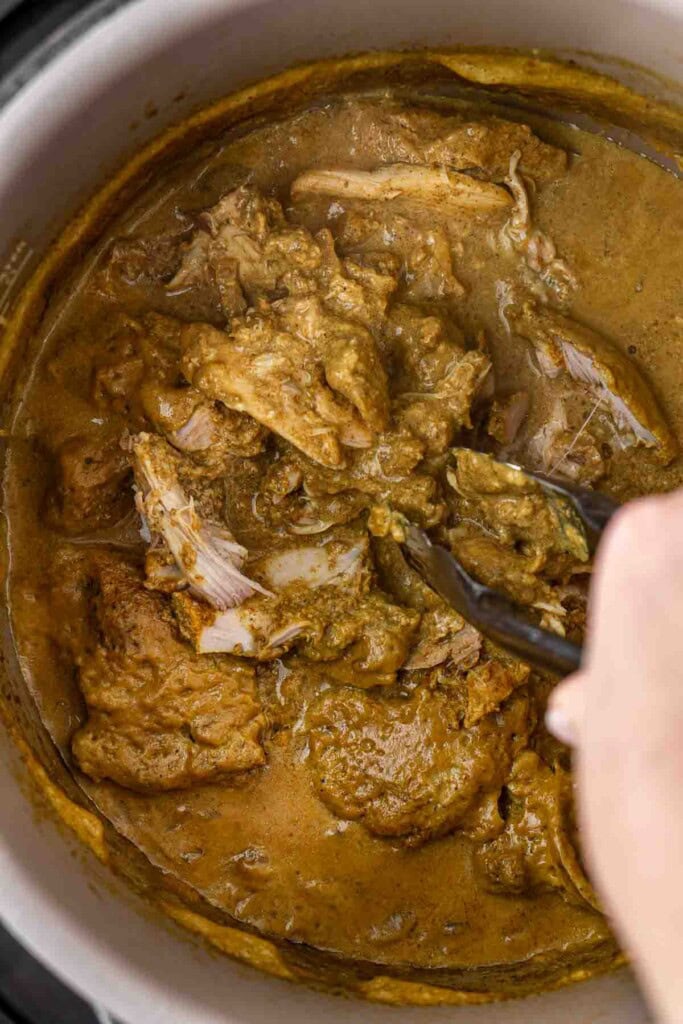 Using tongs to shred the chicken thighs in the slow cooker.