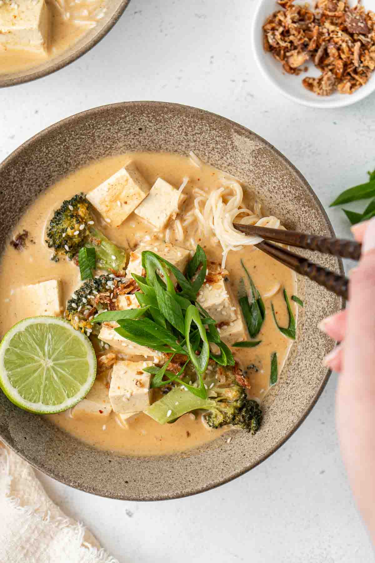 Thai red curry noodle soup in a bowl with spring onions and chopsticks taking a bite of noodles.