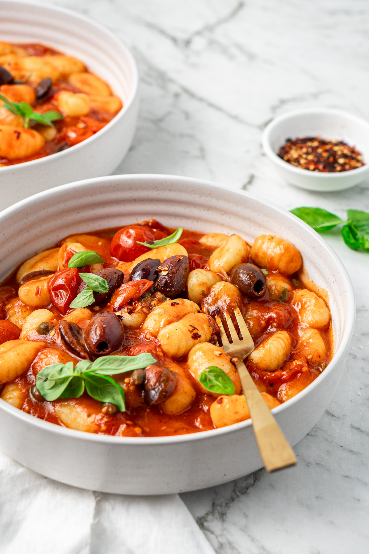 The gnocchi puttanesca in a white bowl with a fork and garnished with fresh basil. 