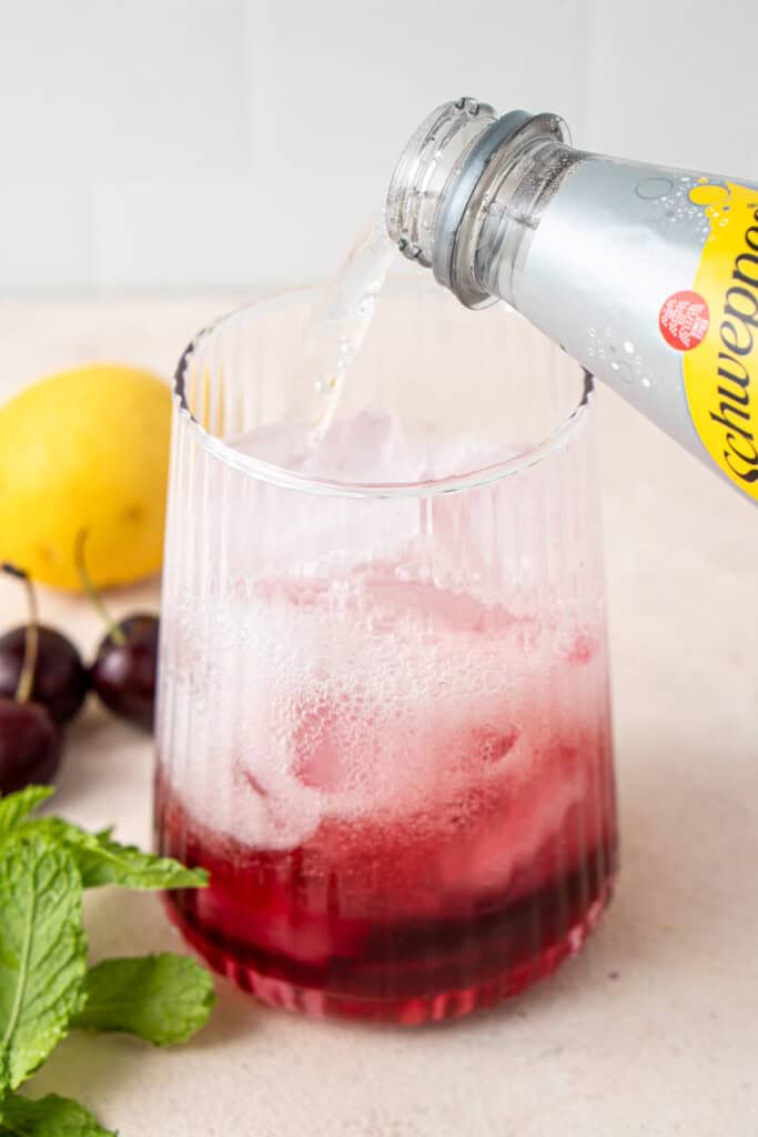 Pouring tonic water into glass over ice and cherry syrup.