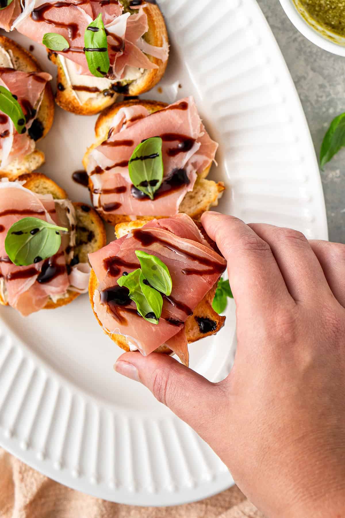 A hand picking up a prosciutto crostini from a serving plate.