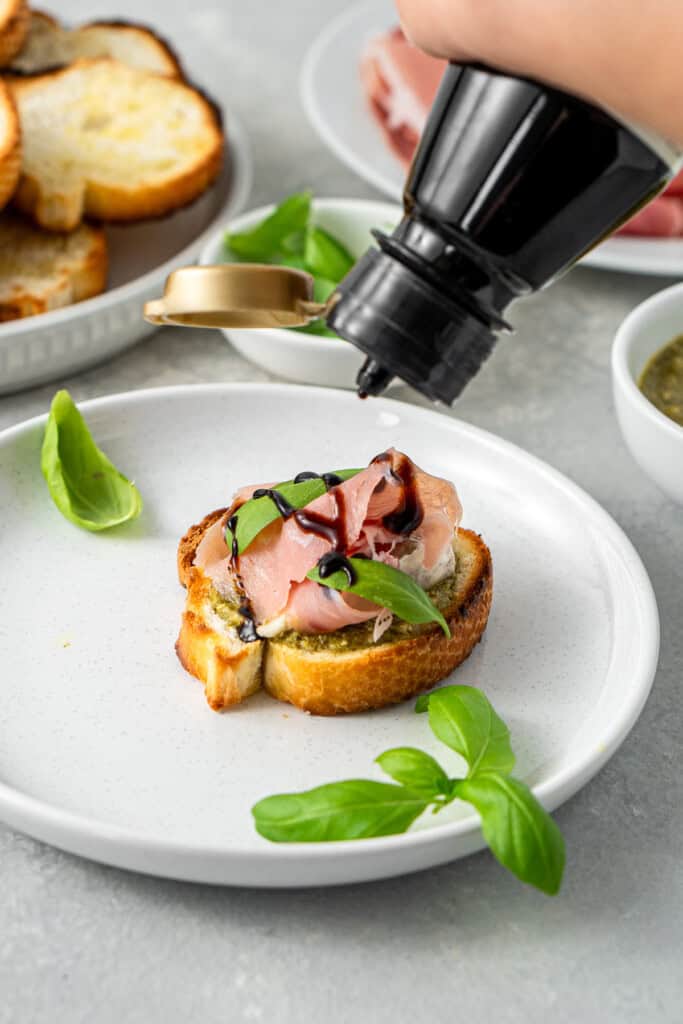 Drizzling balsamic glaze over the prosciutto crostini with basil.