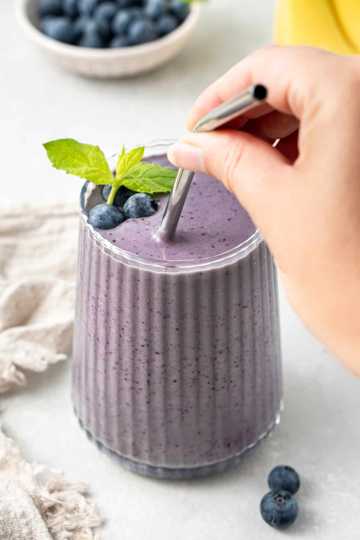 A hand placing a straw into the glass of blueberry banana smoothie.