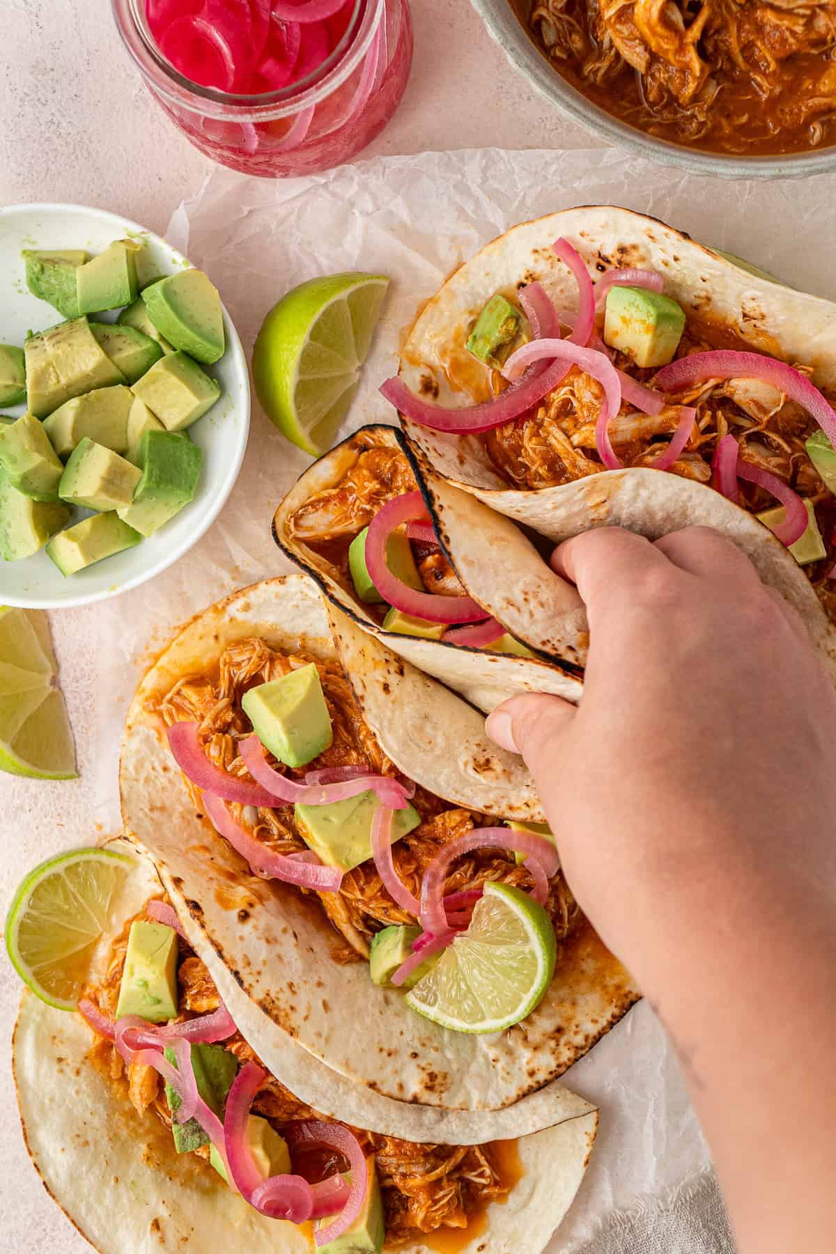 A hand picking up an assembled chicken tinga taco with picked onions, lime and avocado.