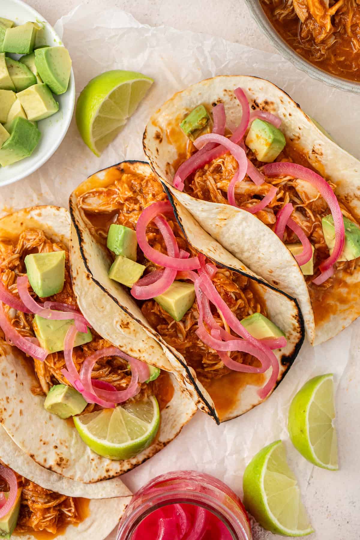 Chicken tinga tacos with avocado, pink pickled onions and lime wedges.