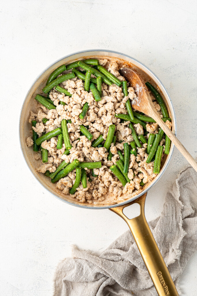 Adding green beans to chicken mince in a frying pan.