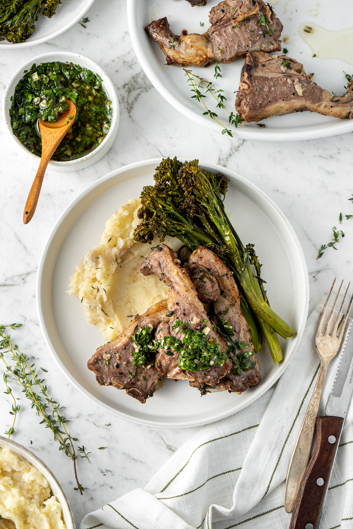 Lamb chops served on a plate with mashed potato, roasted broccolini and chimichurri sauce. 