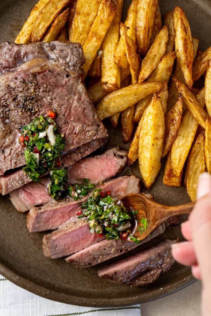 Adding chimichurri to sliced steak on a plate with chips.