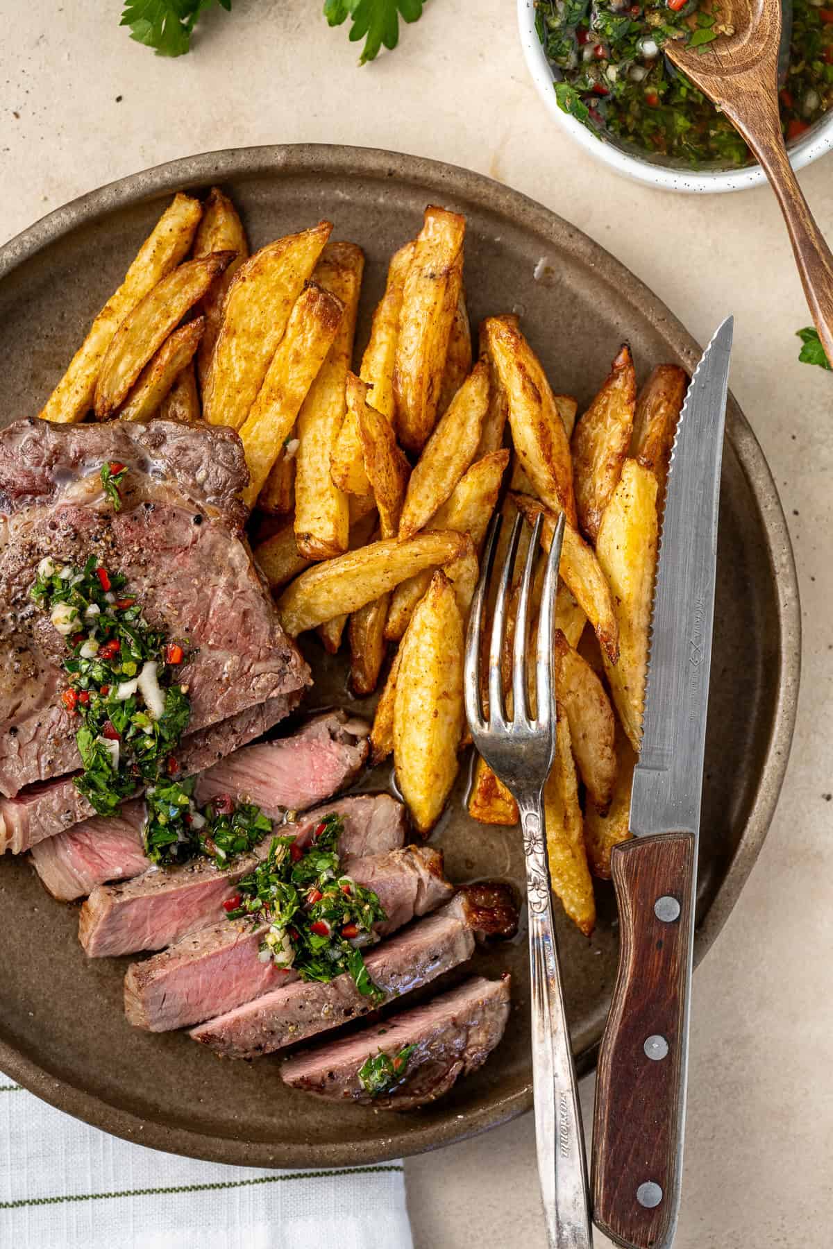 Sliced steak with chimichurri on a plate with chips and a knife and fork.