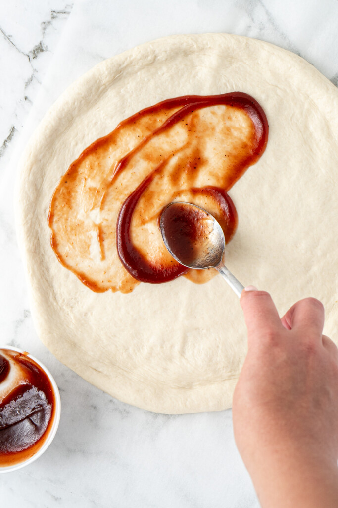 Spreading BBQ sauce onto a homemade pizza base.