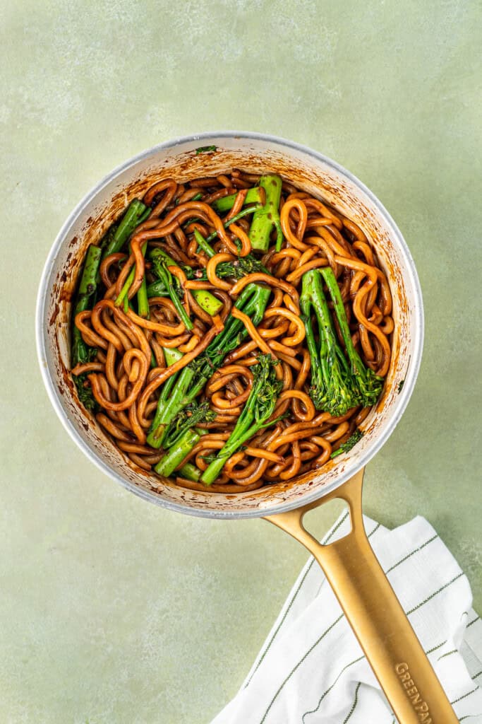 The chilli garlic udon noodles with broccolini in a frying pan.