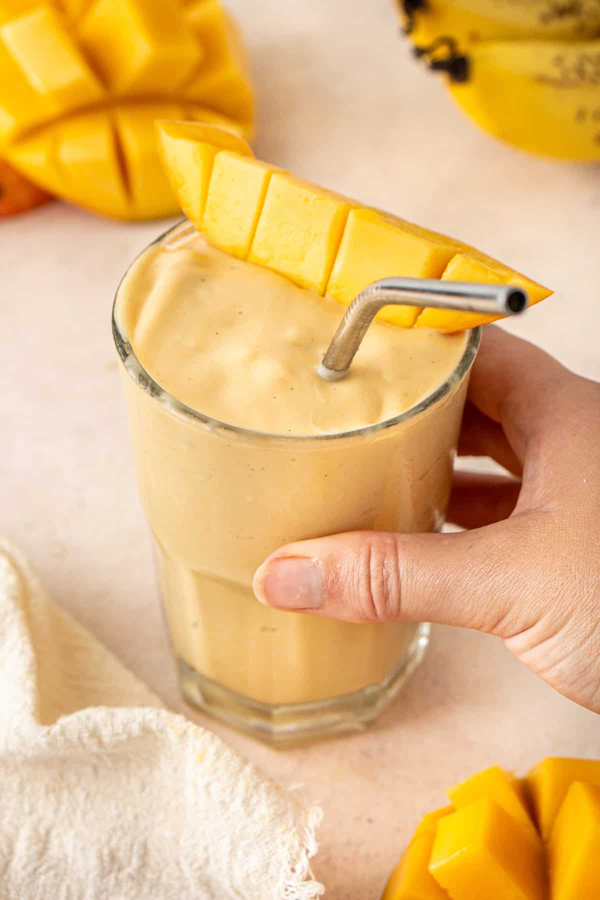 A hand picking up a mango smoothie in a glass with a straw.