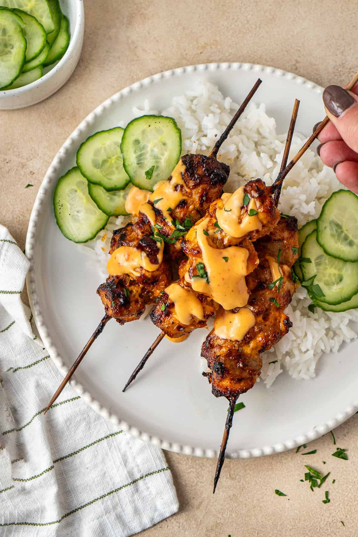 A hand picking up a skewer from a plate with rice and pickled cucumber. 