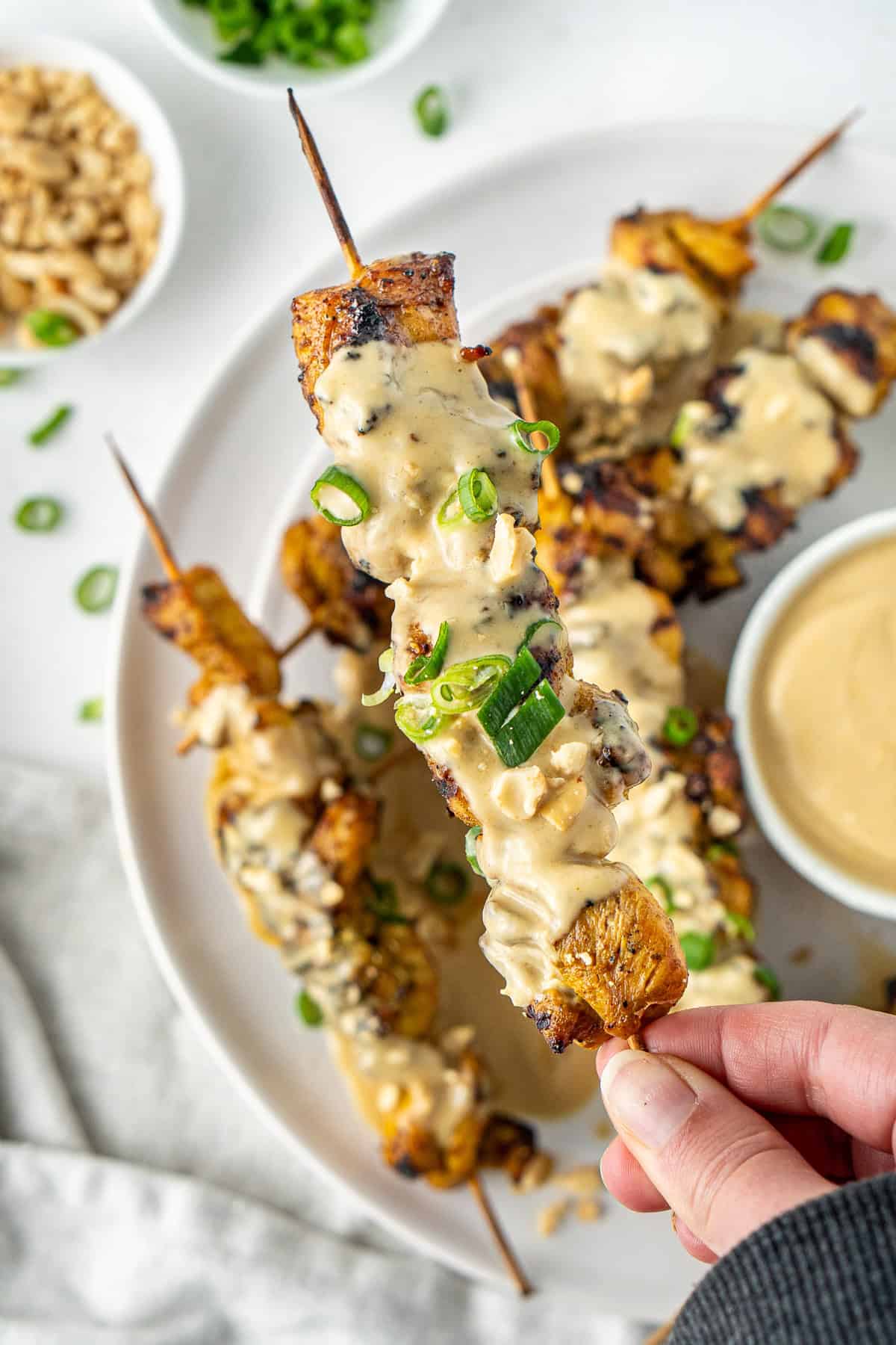 A hand holding a satay chicken skewer coated in peanut sauce. 