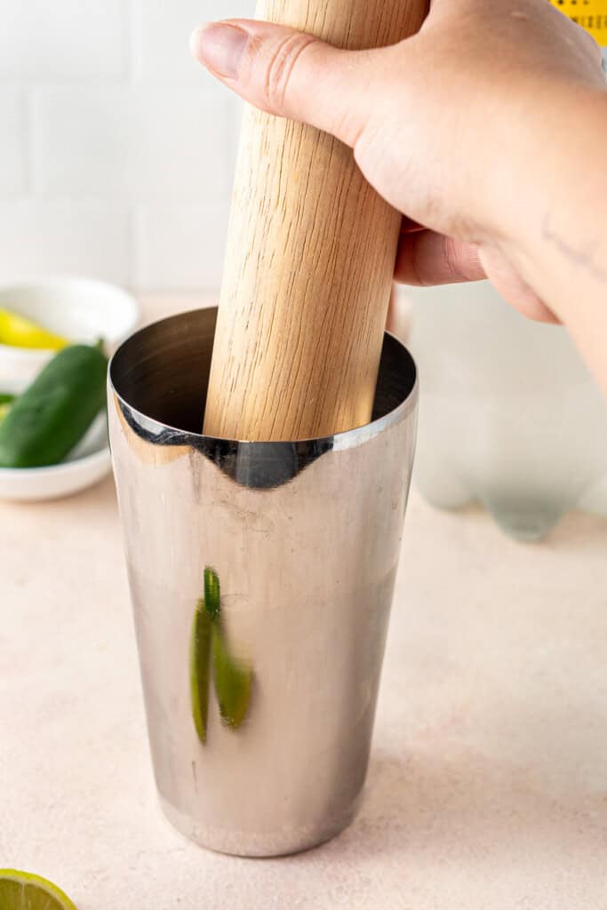 Muddling the jalapeño slices in a cocktail shaker with a wooden rolling pin.