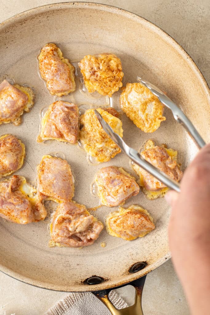 Using tongs to flip the chicken pieces in the frying pan.